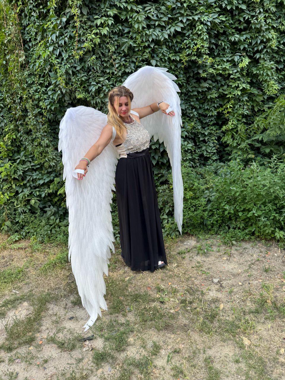 May include: A person wearing a white and black outfit with large white angel wings. The wings are made of layered feathers. The person is standing in front of a green leafy background.