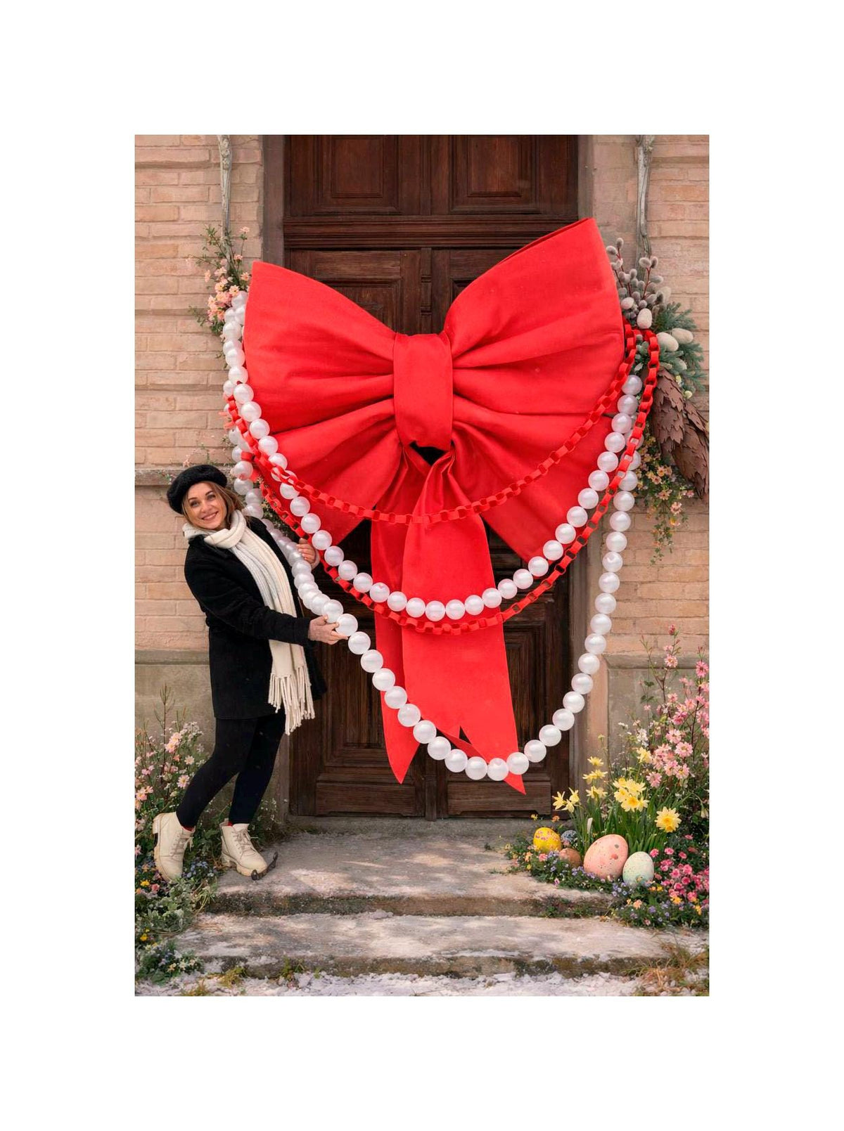 May include: A large, vibrant red bow adorns a wooden door, complemented by a white beaded garland. The festive decoration is surrounded by floral arrangements and a brick facade, creating a welcoming entrance.