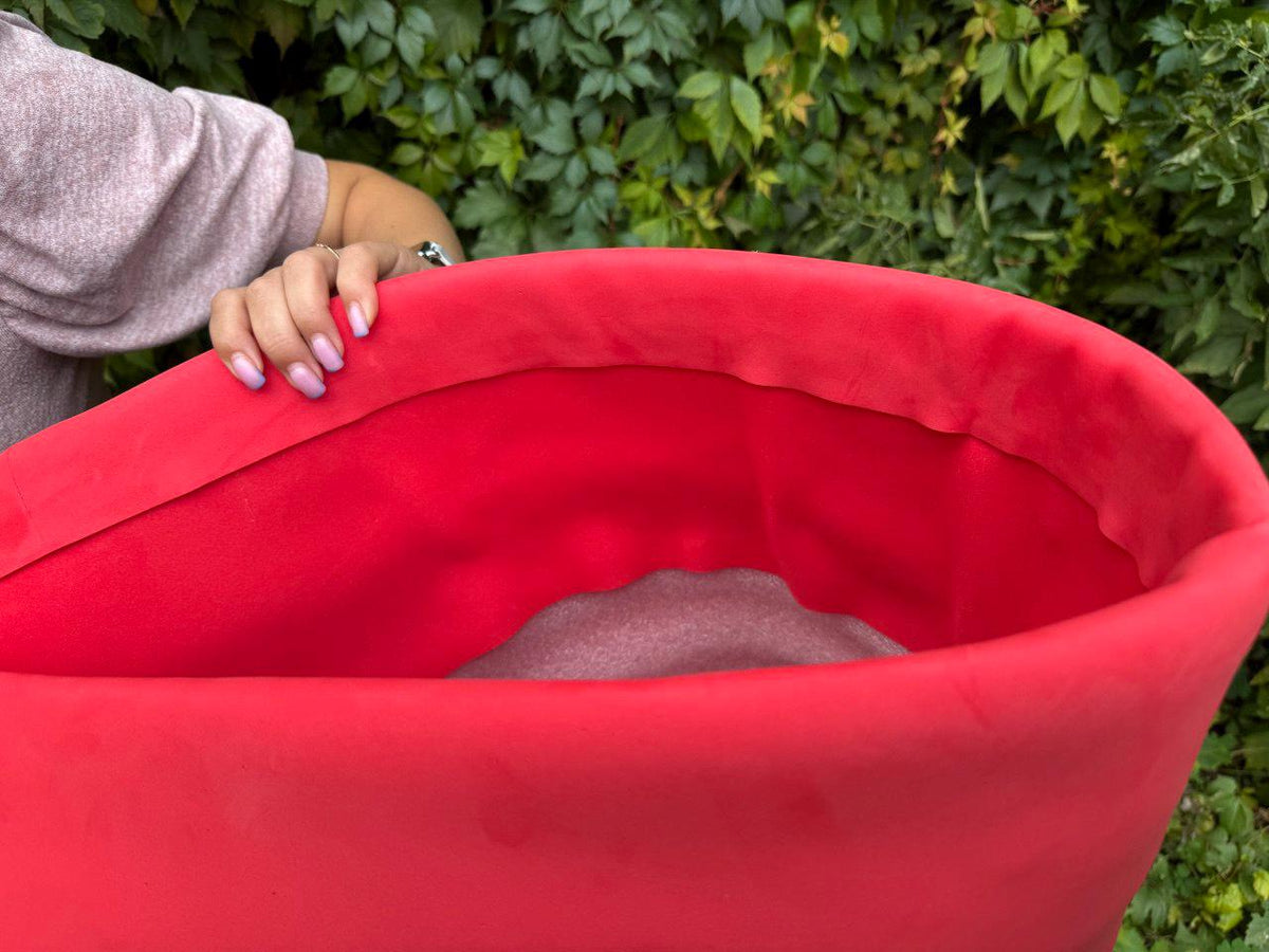May include: A bright red, open-topped container with a soft, velvety texture. The interior is a lighter shade of red. The container is held by a person with a light-colored top and manicured nails. Green foliage is in the background.