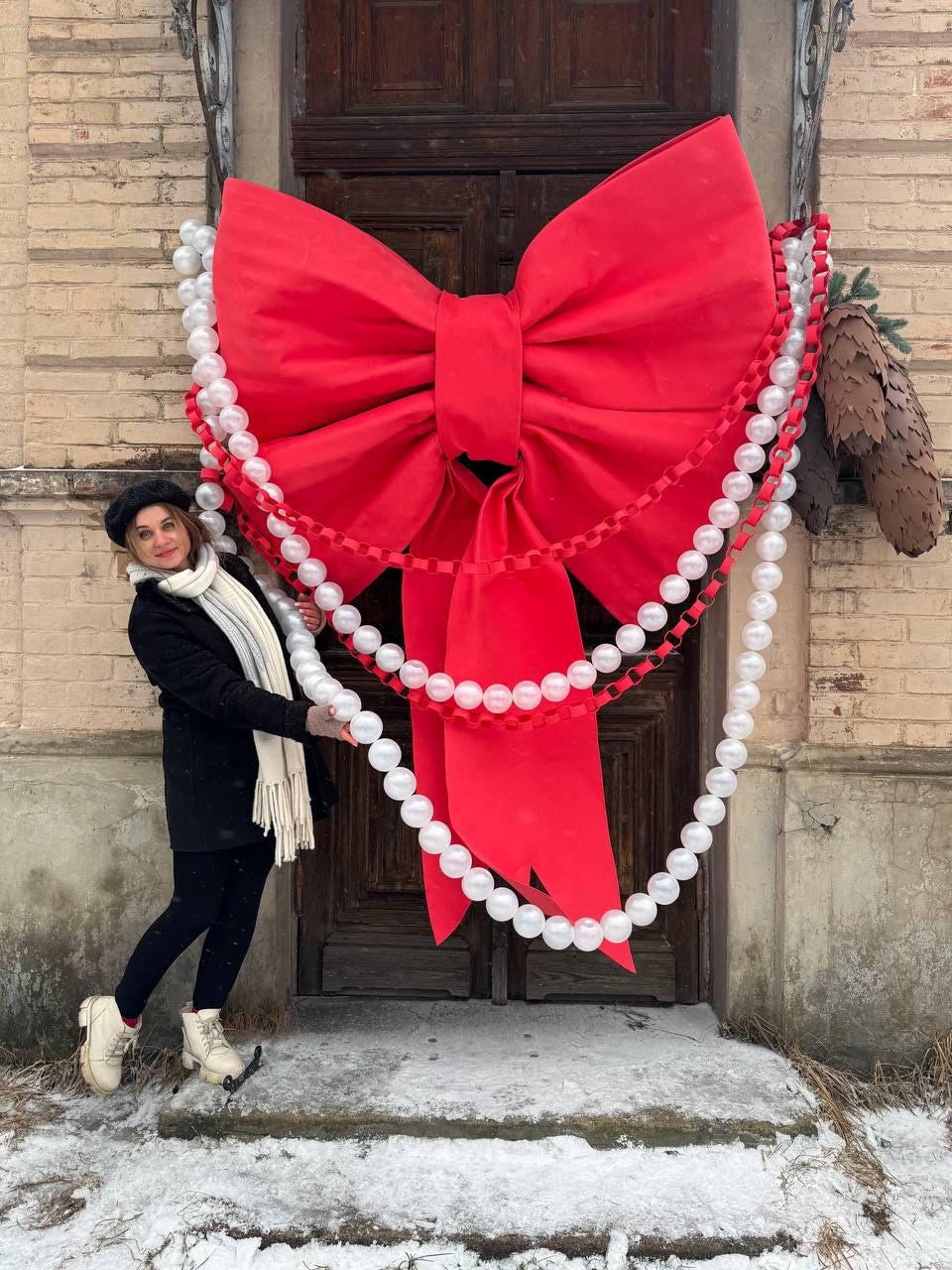 May include: A large, vibrant red bow adorns a wooden door, complemented by strings of white pearl-like beads. The festive decoration is a striking holiday or special occasion statement piece. The bow's size and color create a bold visual impact.