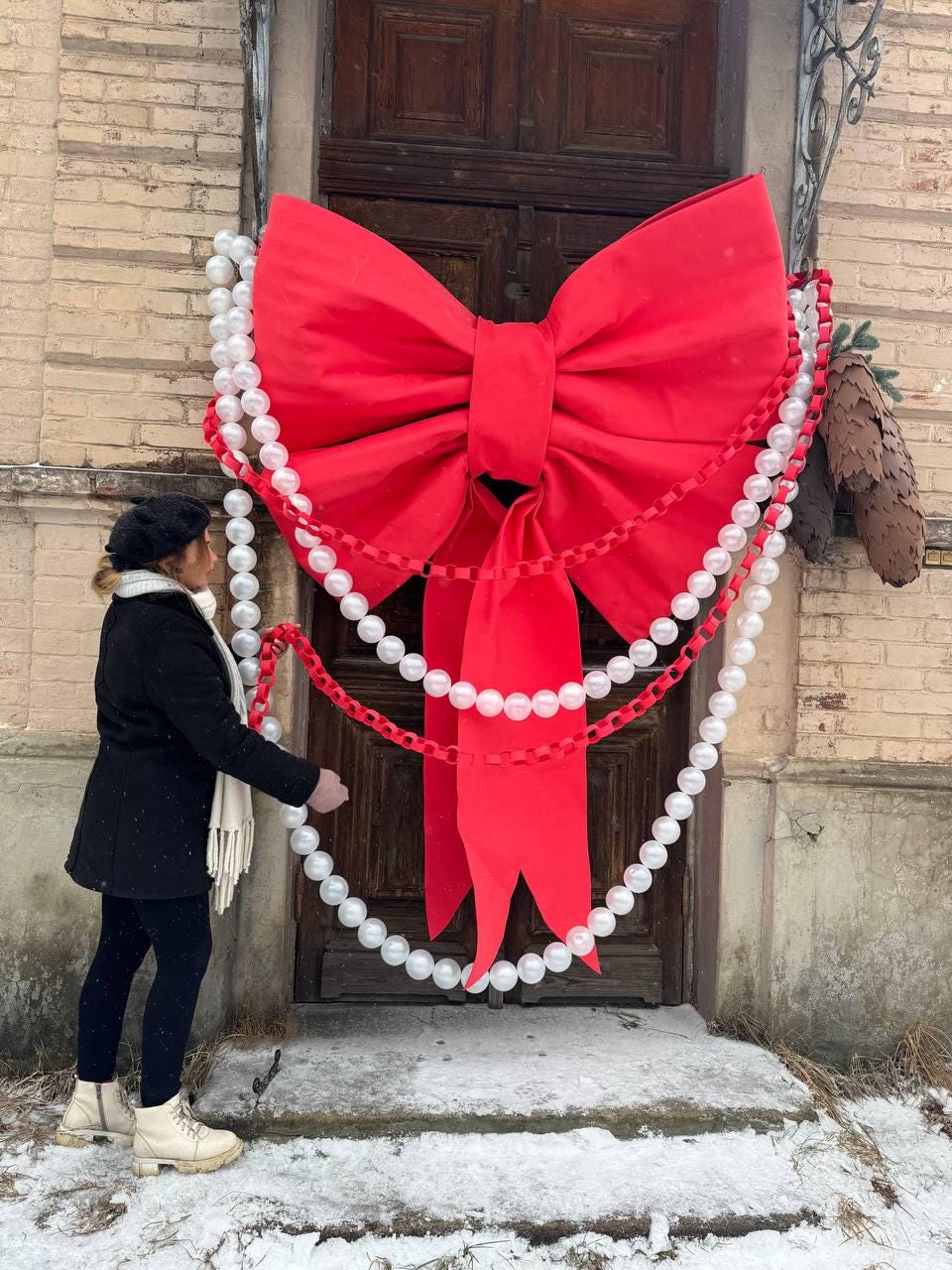 May include: A large, bright red bow adorns a wooden door, complemented by strings of white pearl-like beads and red paper chain. The festive decoration is set against a backdrop of a brick wall and snow-covered ground, creating a holiday or celebratory theme.