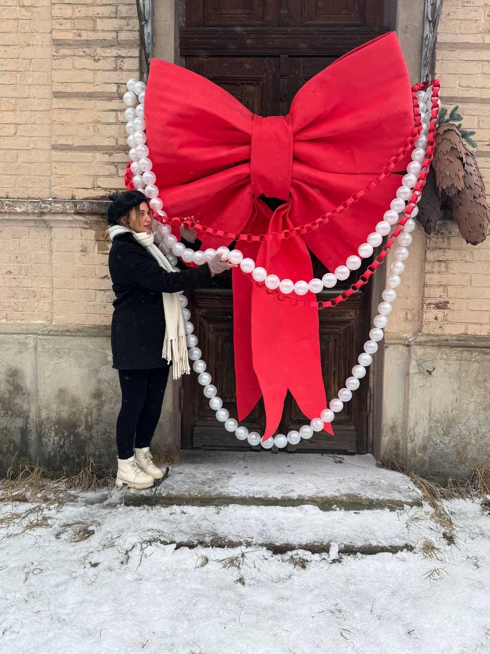 May include: A large, bright red bow adorns a wooden door, complemented by strings of white and red decorative beads. The festive decoration is set against a backdrop of a brick building and a snowy ground.