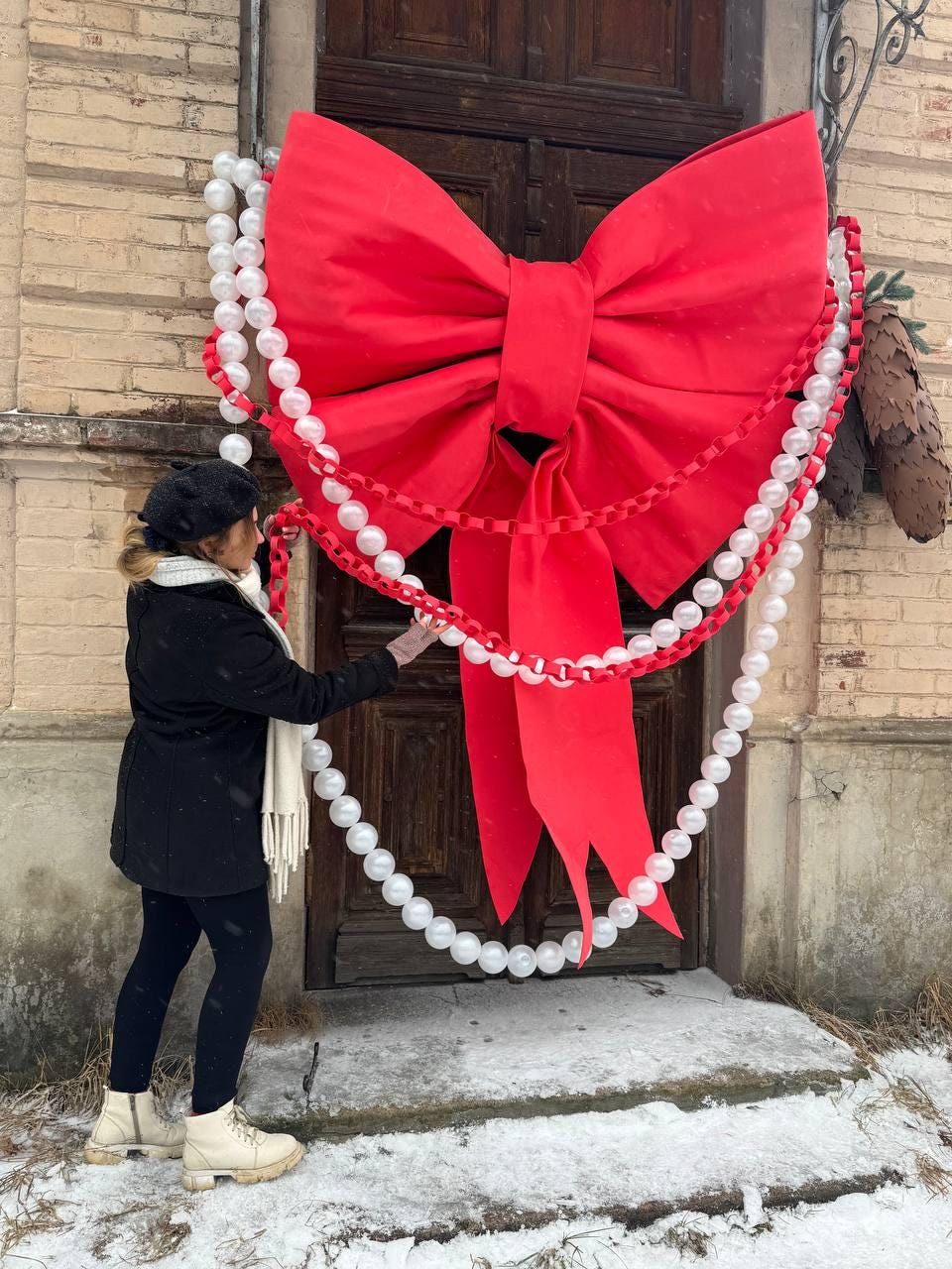 May include: A large, vibrant red bow adorns a wooden door, complemented by strings of white pearl-like beads and red ribbon. The festive decoration suggests a holiday or celebratory theme, adding a touch of elegance to the entrance.