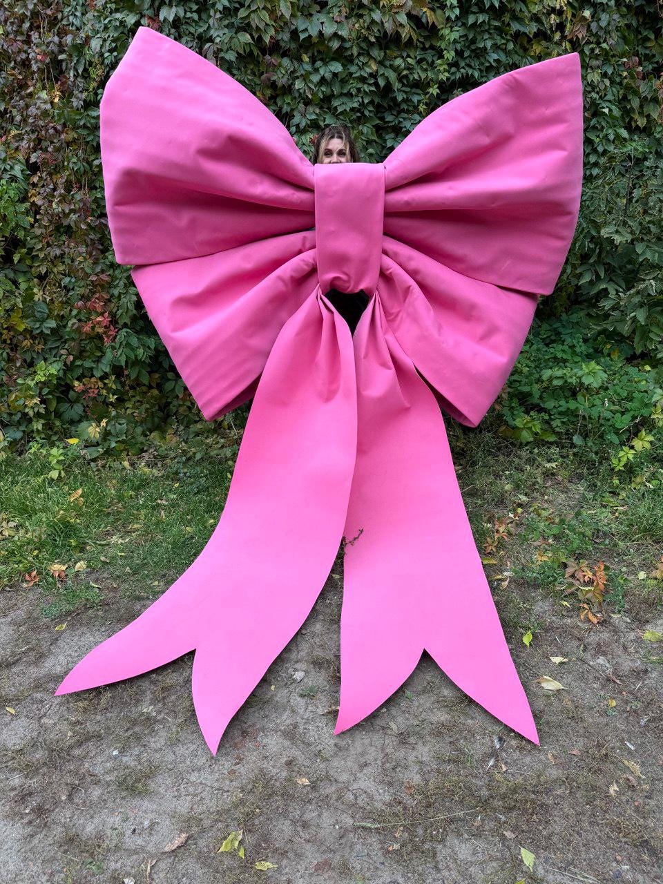 May include: A large, vibrant pink bow with long, flowing tails. The bow is made of a fabric material and is positioned in front of a green leafy background. The bow's size suggests it could be used for decoration or as a prop.