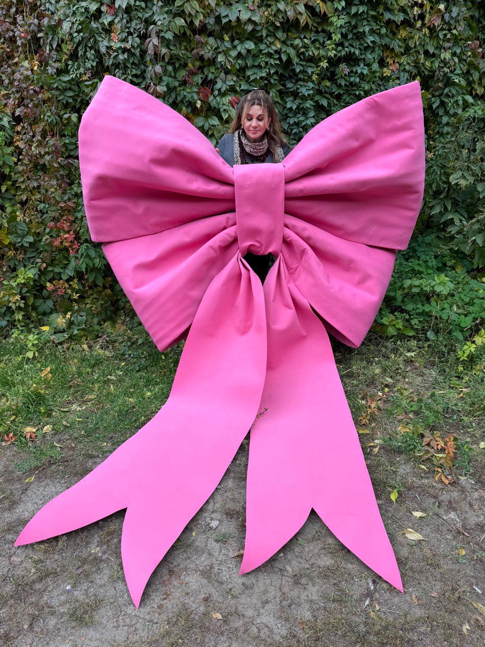 May include: A large, vibrant pink bow with long, flowing ribbons. The bow is made of a smooth, possibly fabric material, and is positioned outdoors against a backdrop of green foliage and grass. The bow's size suggests it's a decorative element.