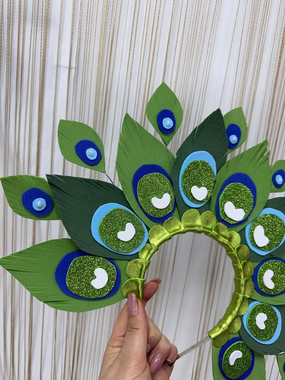 May include: A handmade peacock feather headband. The headband features large green, blue, and glittery green feathers. The feathers are arranged in a fan shape, with a green headband base. The design includes white heart-shaped accents.