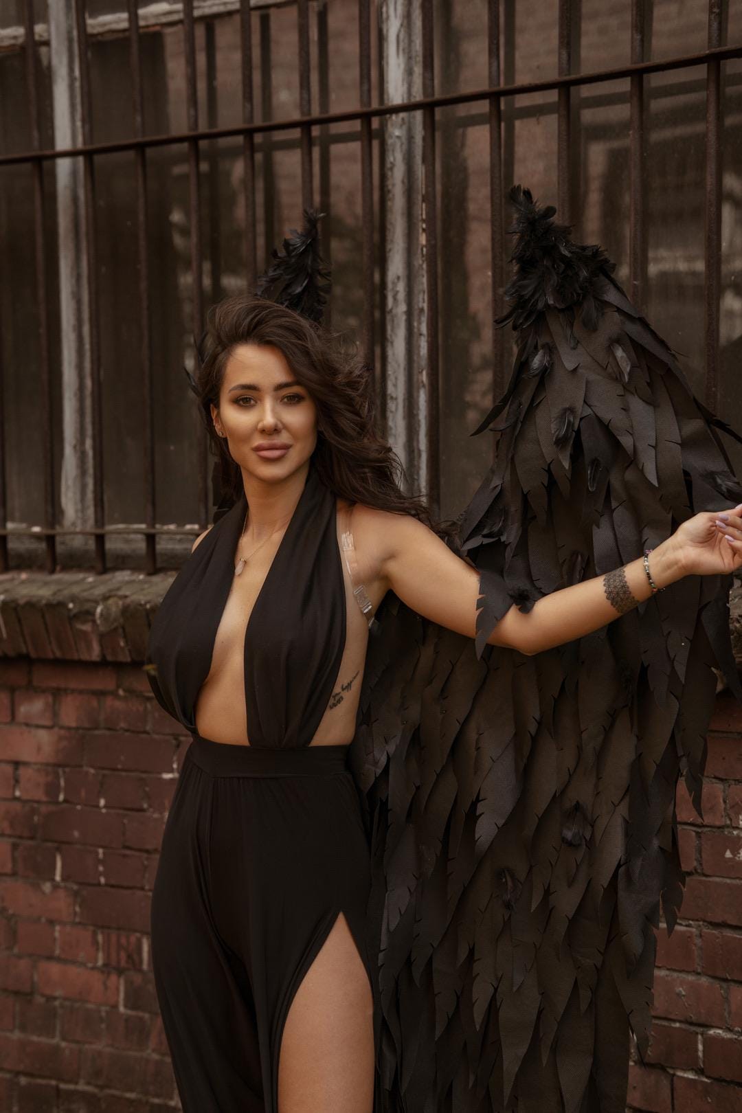 May include: A woman wearing a black outfit and black feathered wings stands in front of a brick wall. The outfit includes a halter top and a skirt with a high slit. The wings are large and detailed.