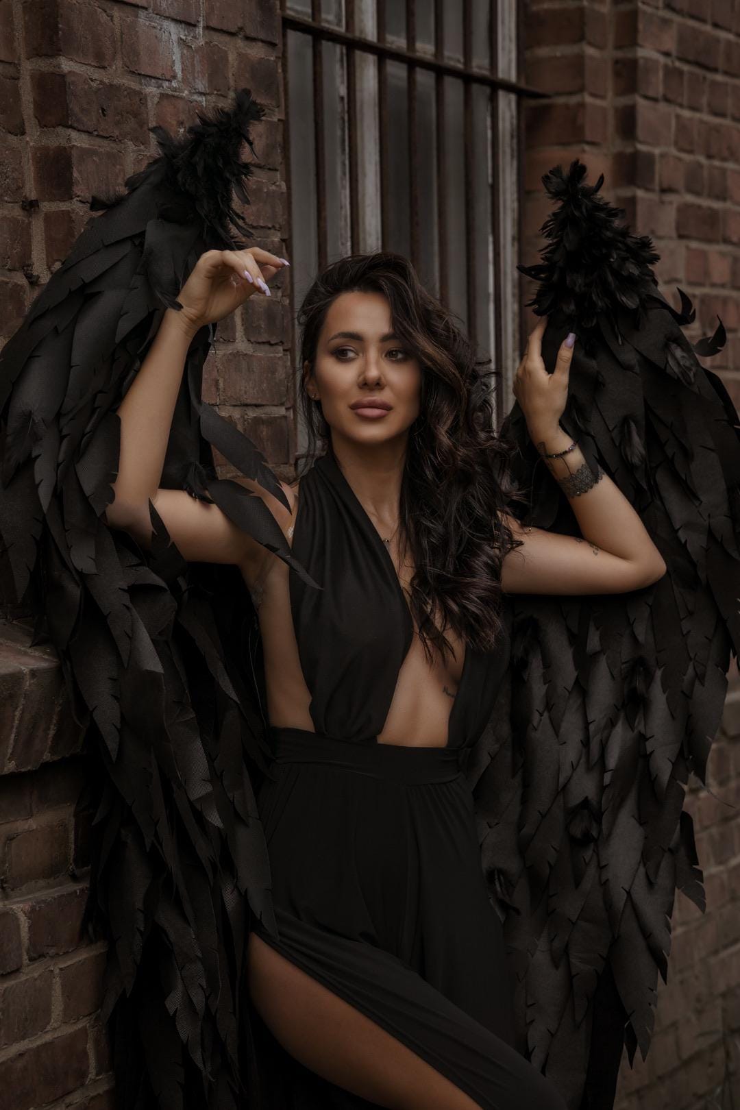 May include: A woman wearing a black dress and large black feathered wings stands against a brick wall. The dress has a plunging neckline and a high slit. The wings are detailed and dramatic, creating a striking contrast against the brick.