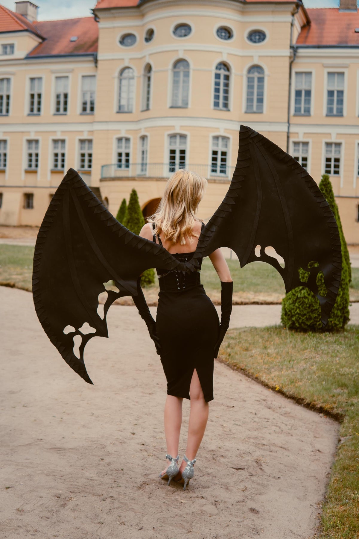 May include: A woman in a black dress and gloves, with large black bat wings, stands on a path in front of a yellow building. The wings have a detailed, textured design with cut-out shapes. The overall aesthetic suggests a costume or cosplay theme.