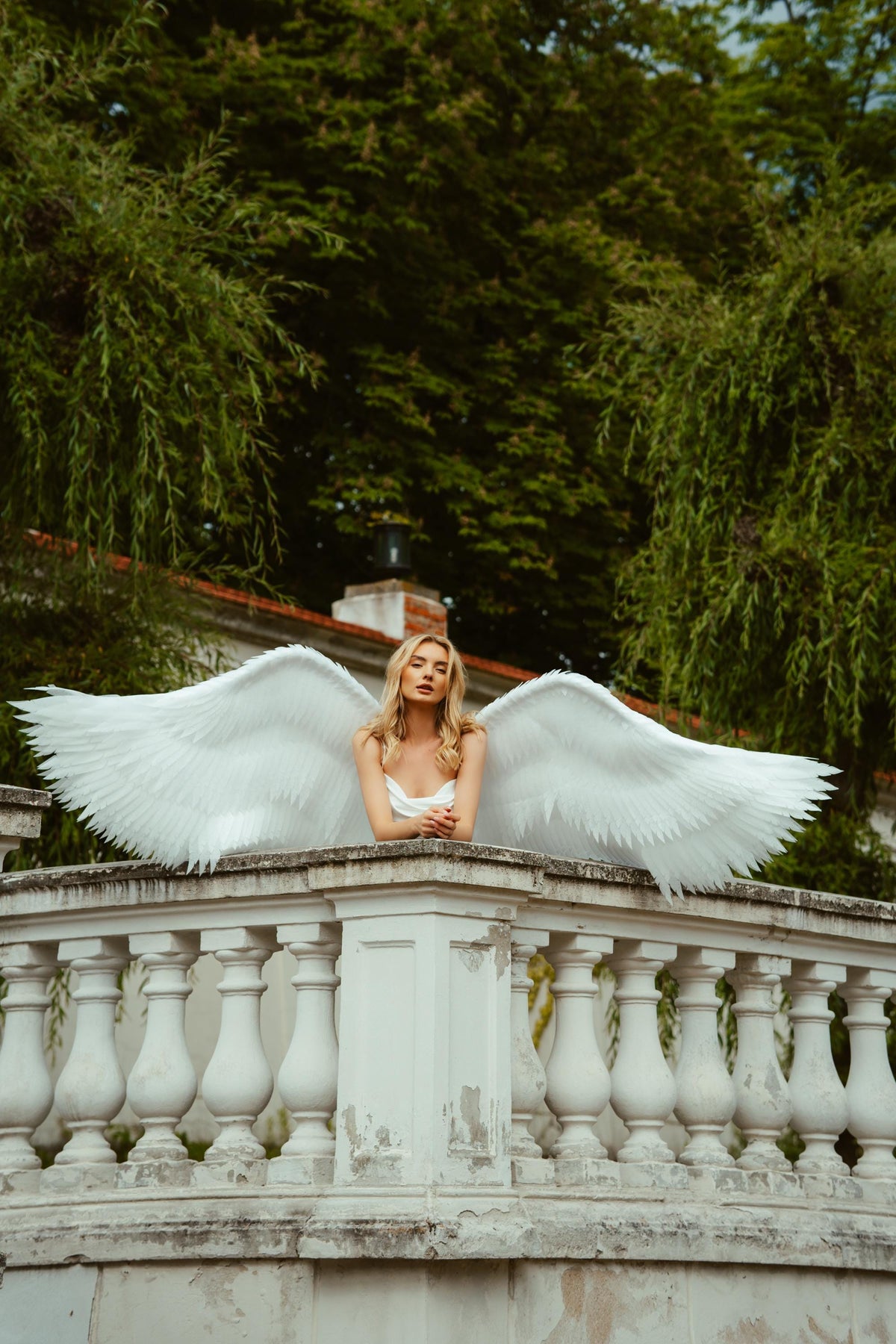 May include: A woman with long blonde hair wearing a white dress and large white feathered angel wings, leaning on a white stone balustrade. The background features lush green trees and a building with a red roof.