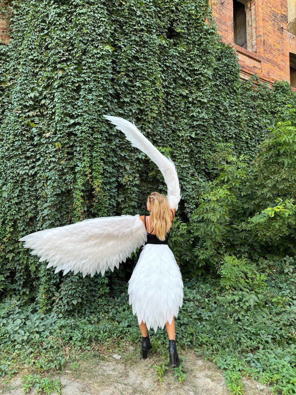 May include: A person wearing a white feathered skirt and matching large white feathered wings. The person is wearing a black top and black boots. The background features lush green foliage and a brick building.