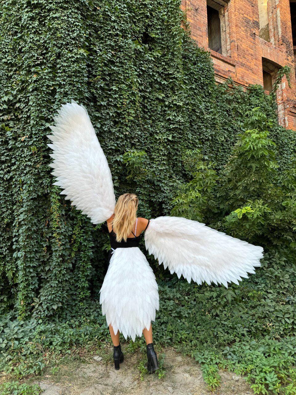 May include: A person wearing a black top and white skirt with large, white feathered angel wings. The person is standing in front of a wall covered in green ivy, with a brick building in the background.