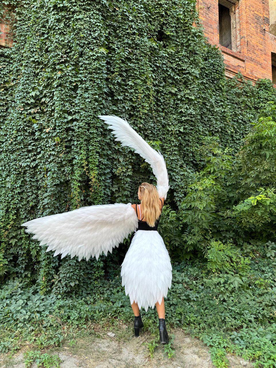 May include: A person wearing a white feathered skirt and large white feathered wings, standing in front of a wall covered in green foliage. The person is wearing a black top and black boots.