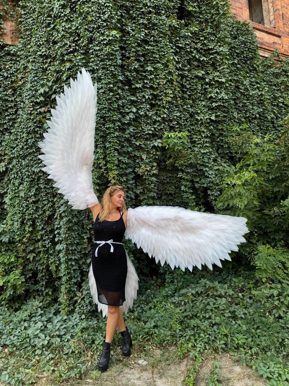 May include: A person wearing large, white angel wings stands in front of a wall covered in green ivy. The wings are made of white feathers and are spread wide. The person is wearing a black dress and black boots.