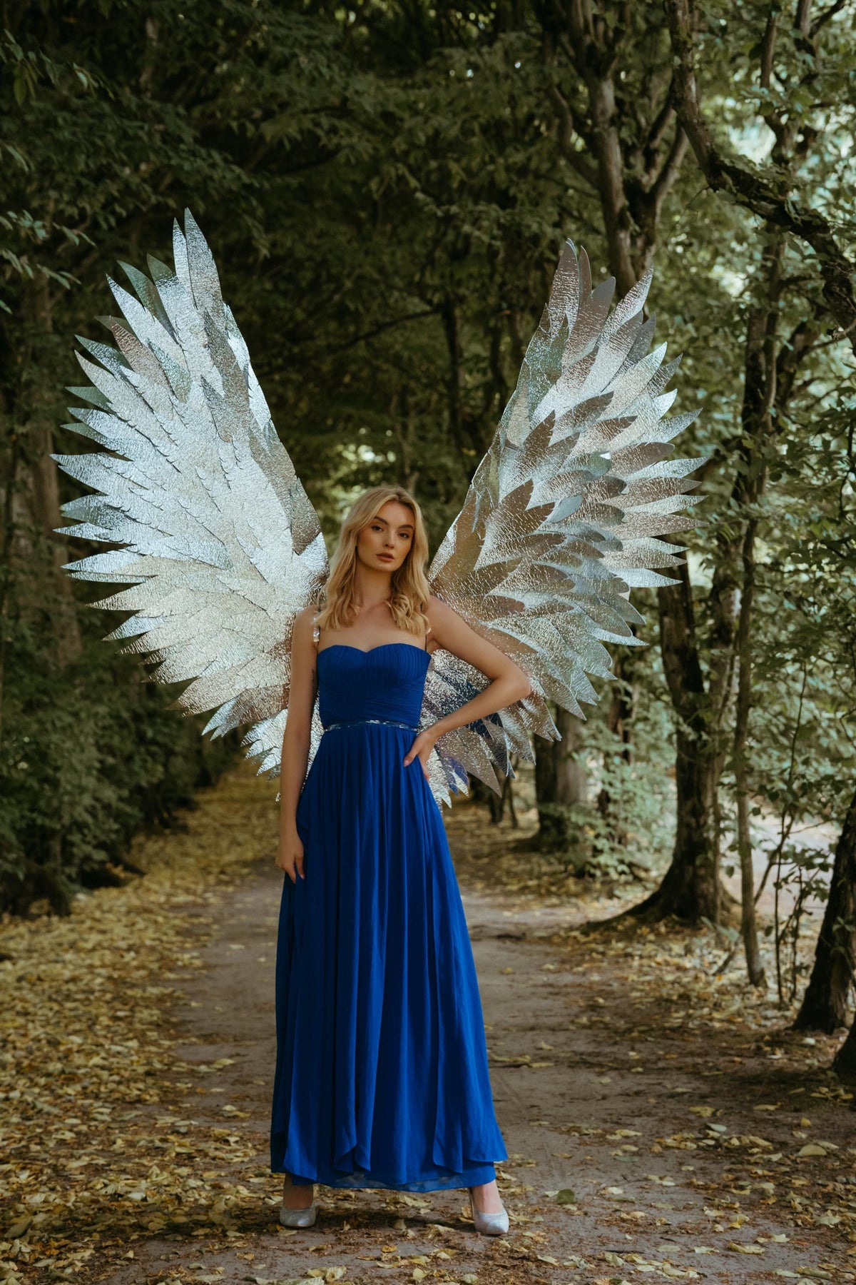 May include: A woman wearing a long, royal blue strapless gown and silver angel wings stands on a path in a forest. The wings are large and silver, with a layered feather design. The dress has a fitted bodice and a flowing skirt.