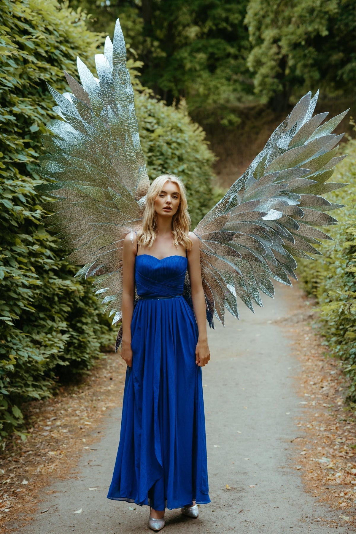 May include: A woman wearing a long, royal blue dress and large silver angel wings stands on a path in a wooded area. The wings are detailed with multiple layers of reflective material. The dress is strapless with a fitted bodice and flowing skirt.