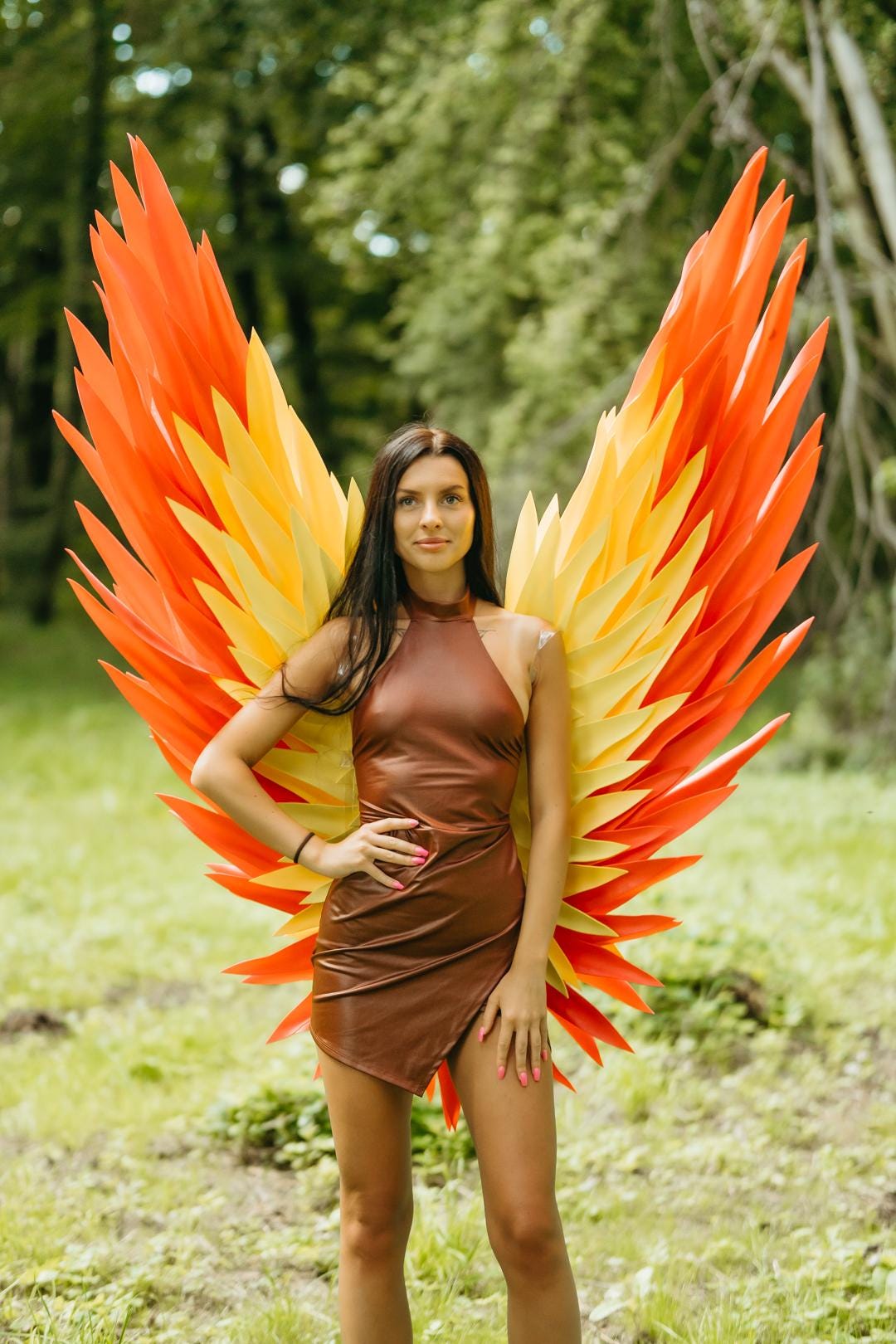 May include: A brown sleeveless mini dress with a high neckline and a wrap-style skirt, paired with large, oversized decorative wings featuring layered, pointed feathers in vibrant shades of bright orange and yellow, extending outward from the back.