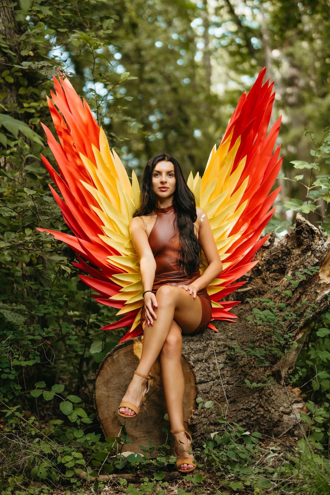 May include: A woman wearing a brown dress and tan heels sits on a tree stump, with large red, yellow, and orange wings behind her. The wings are designed to resemble flames. The background is a lush green forest.