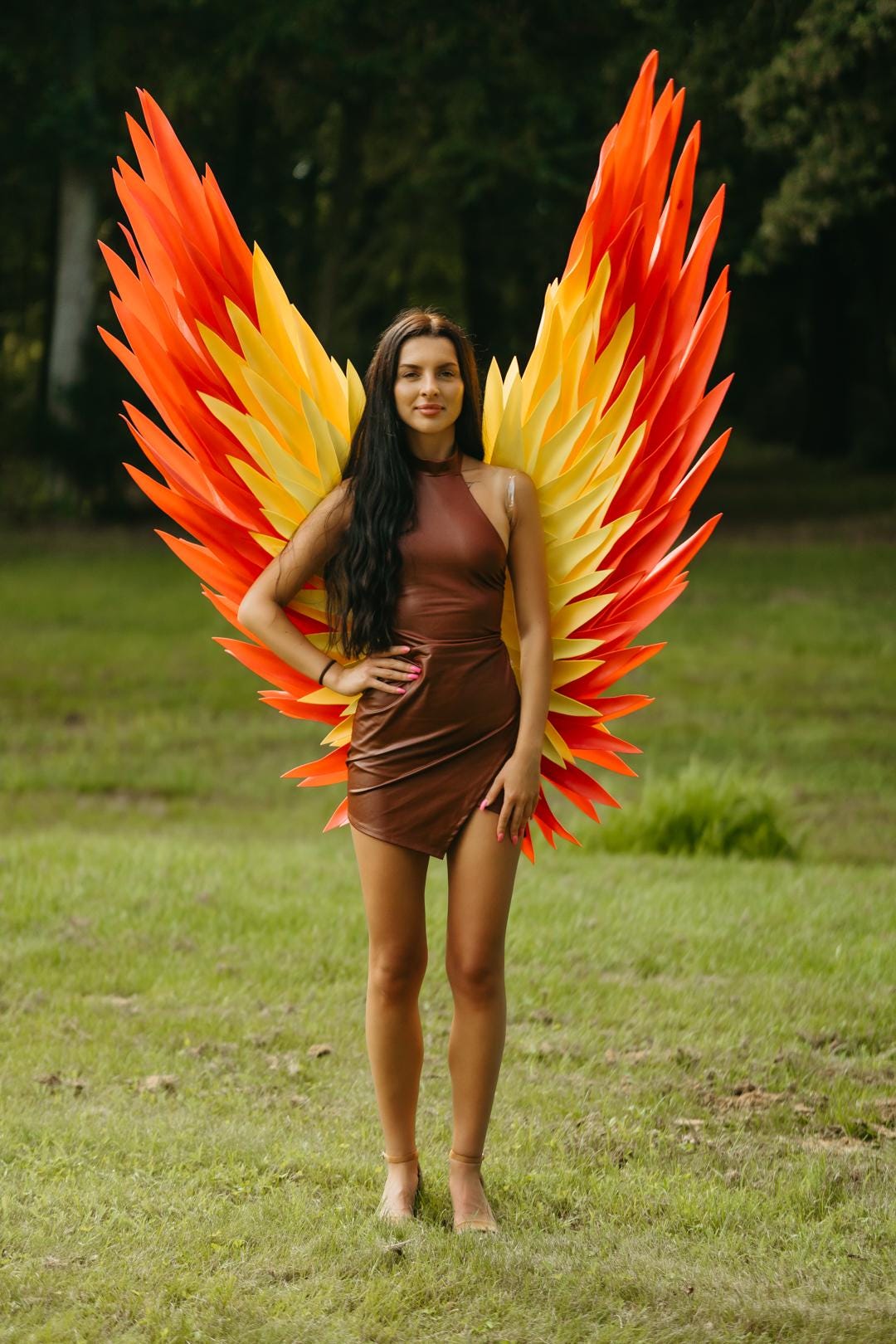 May include: A woman wearing a brown dress and large, colorful wings. The wings are a combination of red, yellow, and orange, resembling flames. The dress is a halter-style with a wrap skirt. The background is a grassy field with trees.