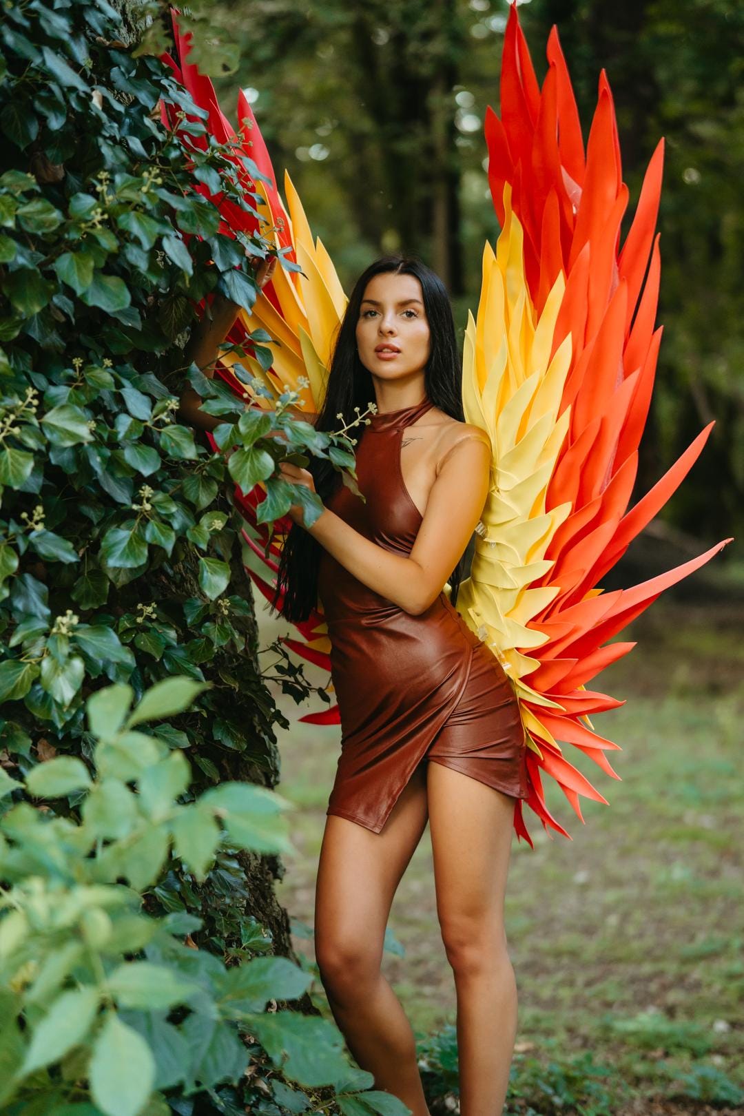 May include: A woman wearing a brown leather dress and large, colorful wings. The wings are a combination of red, yellow, and orange. The dress is a wrap style, and the background is a lush green forest.