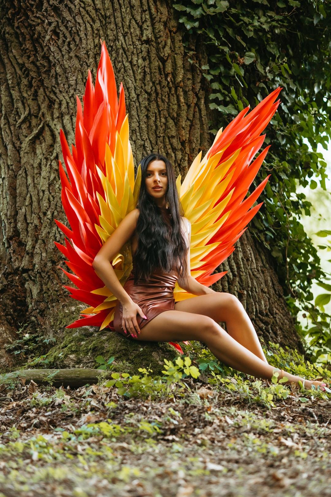 May include: Large decorative wings featuring layered, pointed feathers in vibrant shades of red, orange, and yellow. The wings are structured with a rigid frame and display a gradient color pattern, extending upwards and outwards from the center.