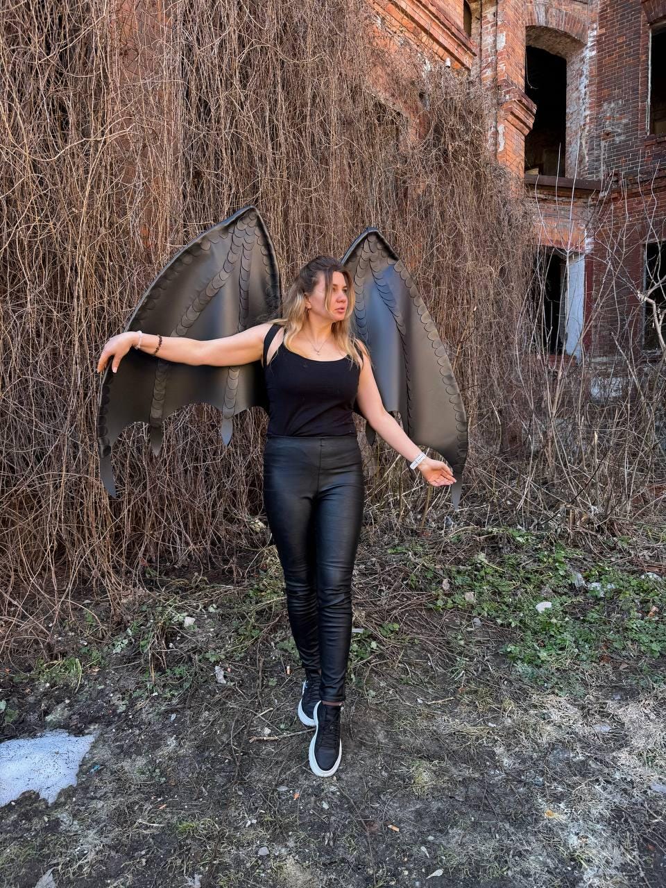 May include: A pair of large, matte black dragon wings with a scalloped edge design, worn as a backpack accessory. The wings feature a structured, layered feather-like pattern and are attached to black straps, extending outward from the shoulders.
