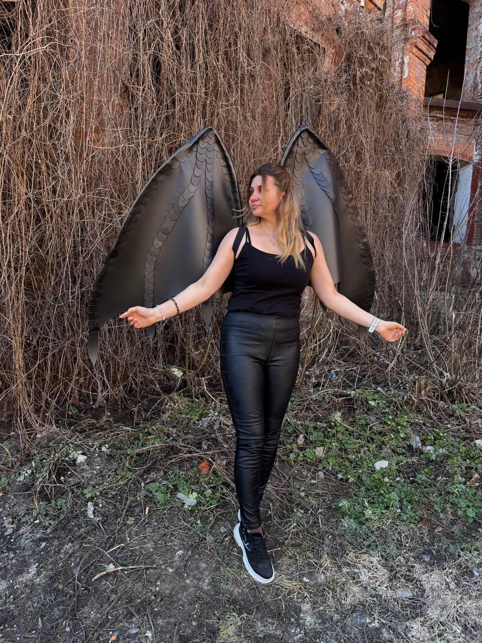 May include: Large black wings with a layered feather-like design attached to a black tank top, paired with black fitted trousers and black sneakers. The wings feature a structured, pointed shape with visible segmented panels and dark trim.