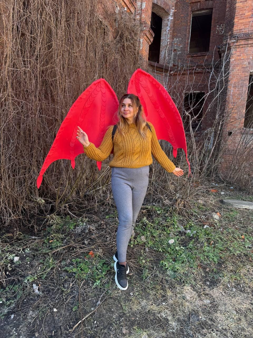May include: A pair of large, bright red fabric wings with a textured, scale-like pattern, worn over a mustard yellow cable-knit sweater, gray leggings, and black sneakers. The wings feature pointed tips and a structured, lightweight design.