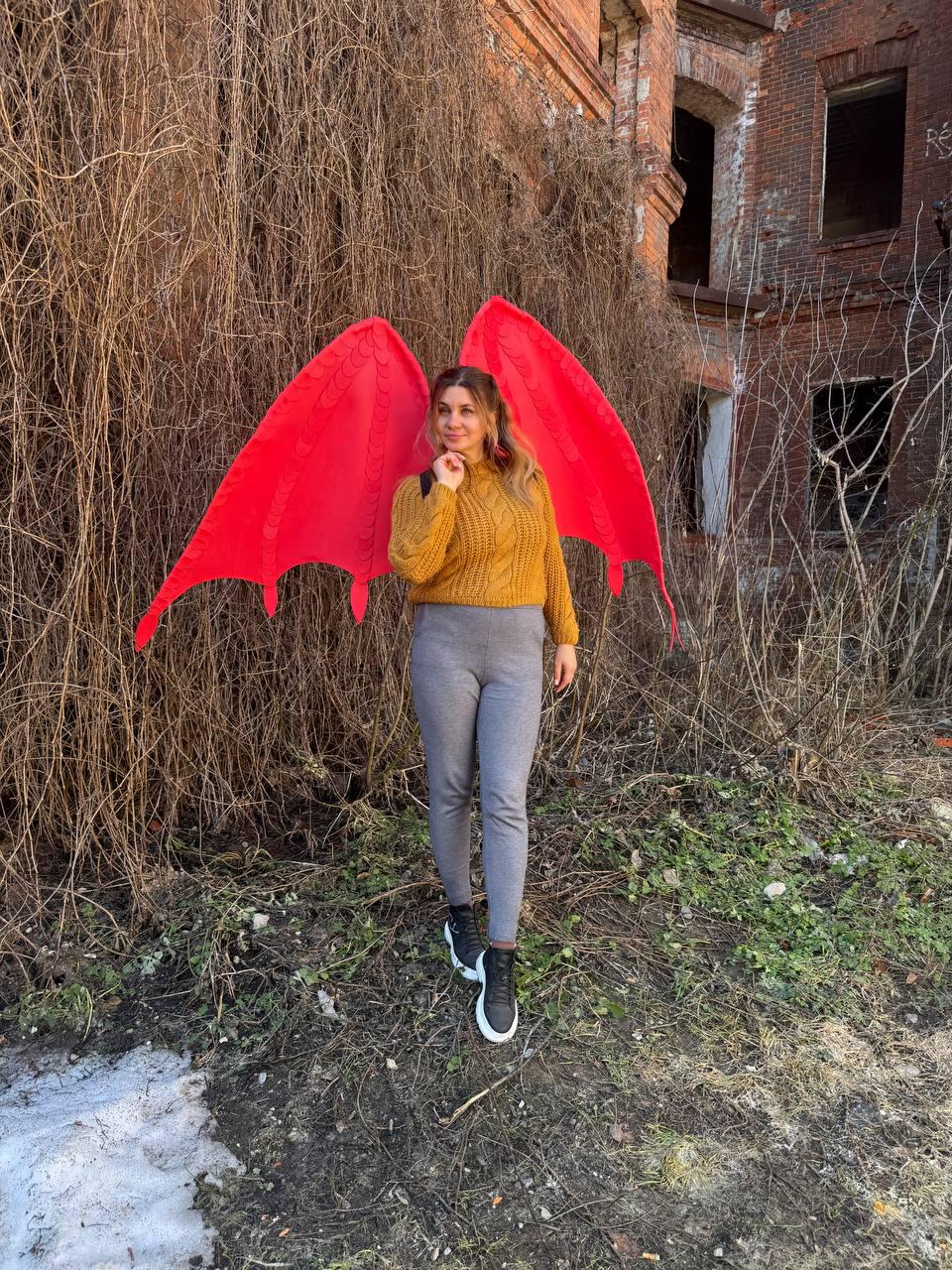May include: A pair of large, vibrant red fabric dragon wings with a structured, pointed design, worn over a mustard yellow cable-knit sweater, grey fitted trousers, and black sneakers with white soles, standing in an outdoor setting.