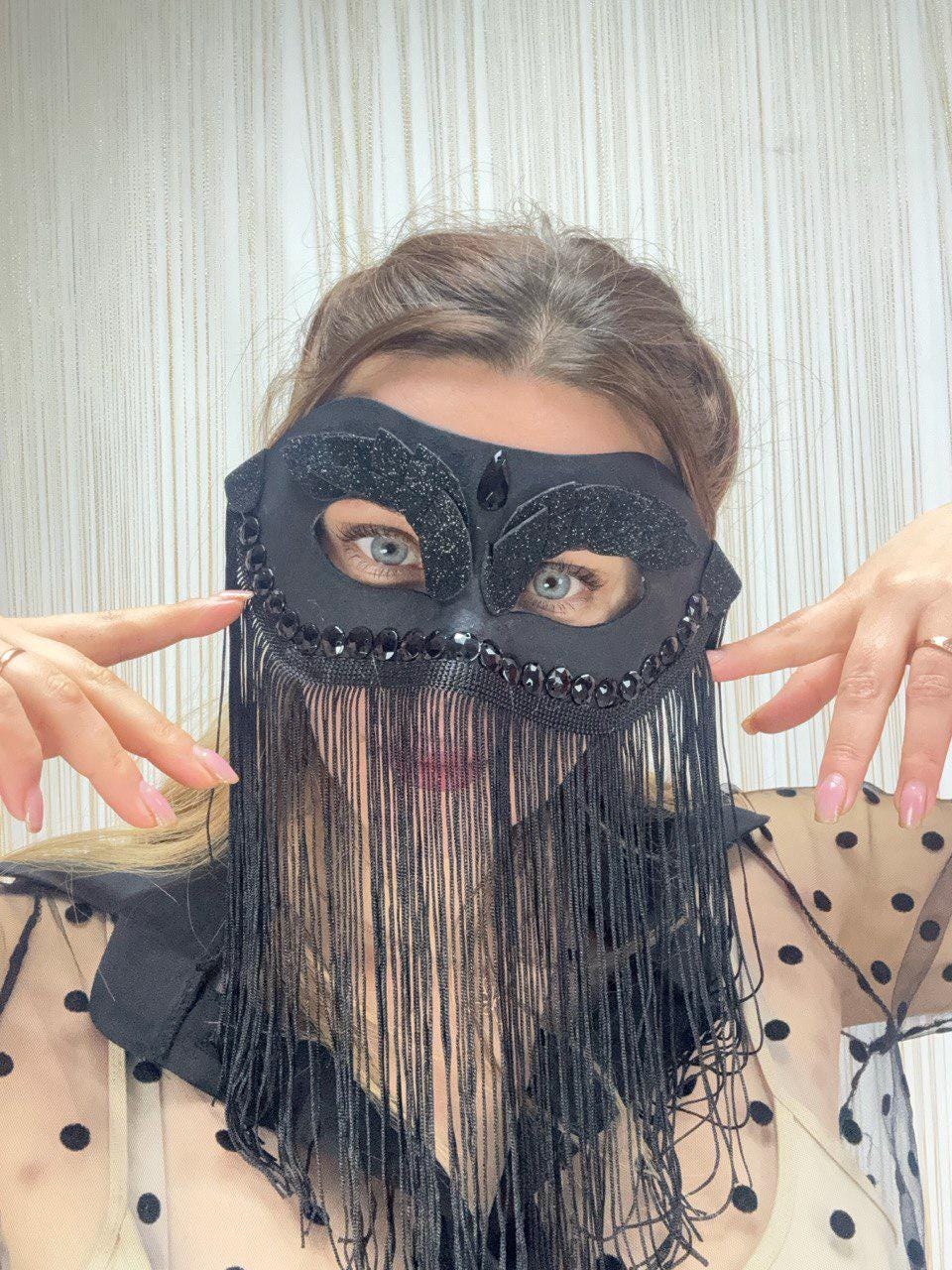 May include: Black masquerade mask with a beaded brow and fringe. The mask has a teardrop-shaped gem above each eye and is decorated with small, dark beads. Long, black fringe hangs from the bottom of the mask.