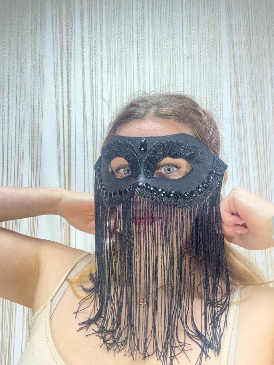 May include: Black masquerade mask with a decorative design and long fringe. The mask features a beaded upper edge and a central jewel. The mask is designed to cover the eyes and upper face.