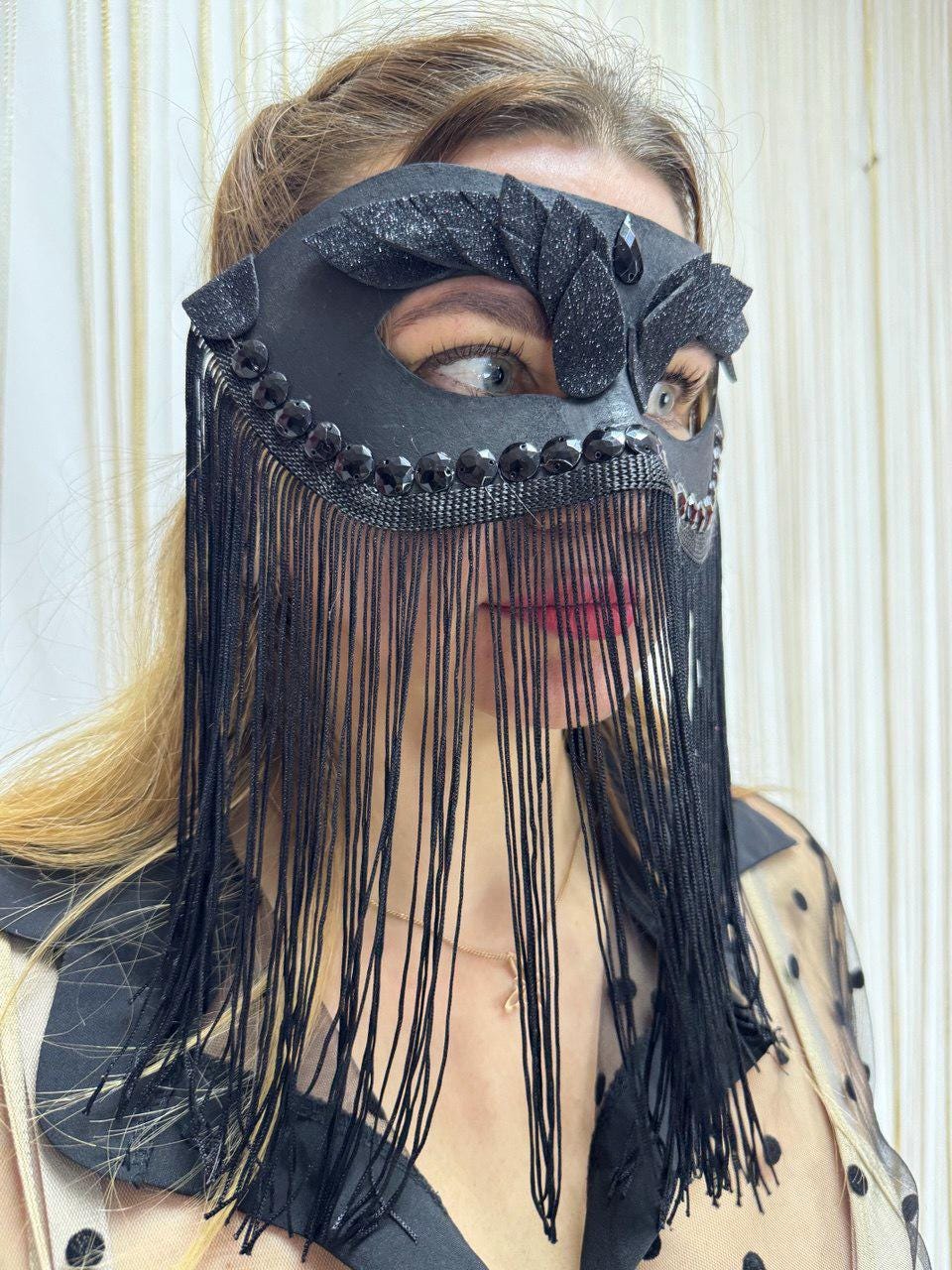 May include: Black masquerade mask with fringe. The mask features a decorative design with black beads and glitter accents. The fringe hangs down, partially obscuring the lower face. The mask is worn by a person.
