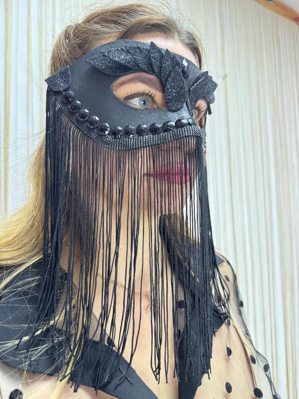 May include: Black masquerade mask with a decorative design and long fringe. The mask features a black base with embellishments and a fringe that hangs down. The mask is designed to cover the eyes and upper face.