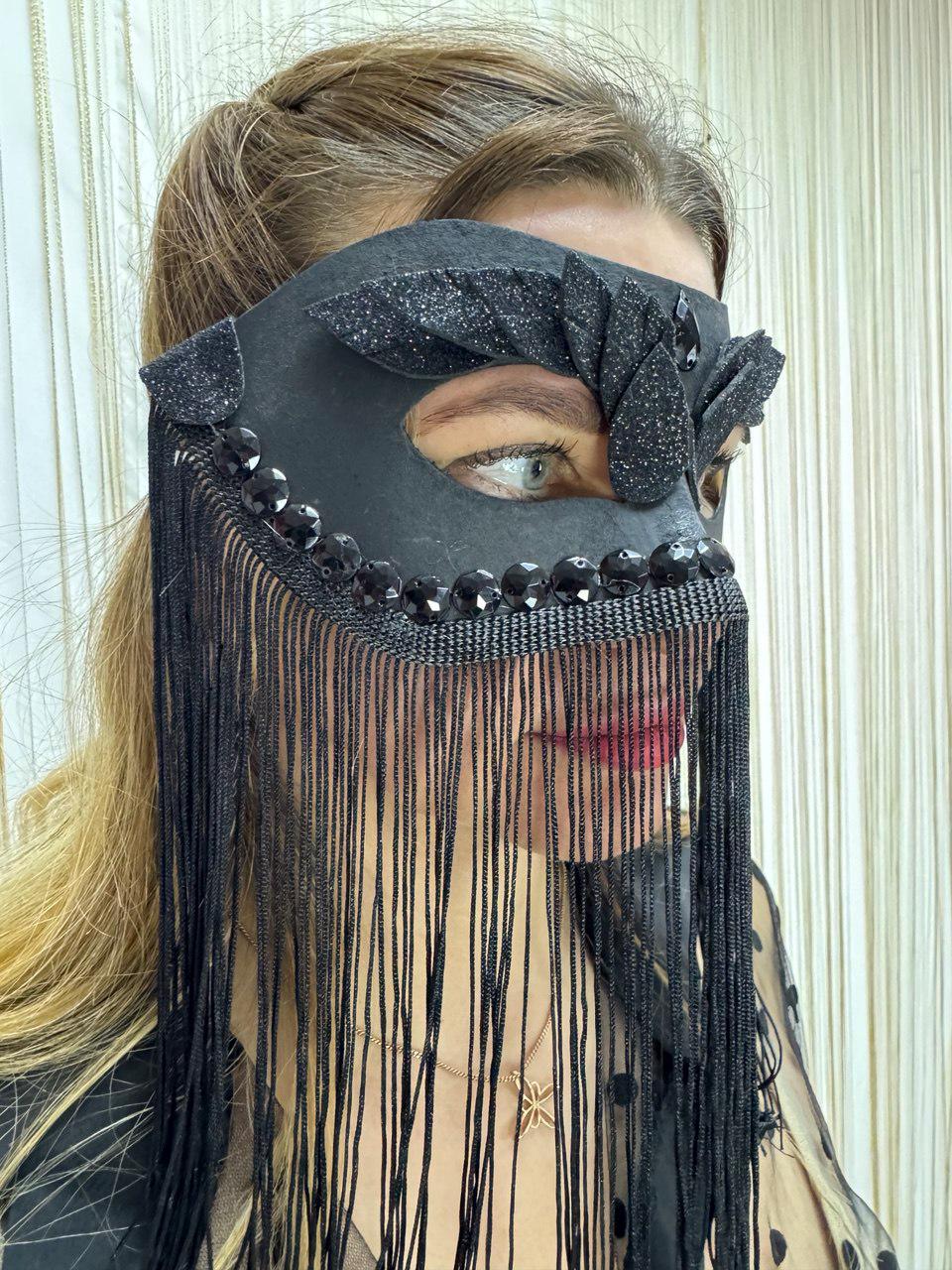 May include: Black masquerade mask with beaded accents and fringe. The mask features a floral design with glitter detailing and is adorned with black beads along the top and fringe. The fringe hangs down, partially obscuring the lower face.
