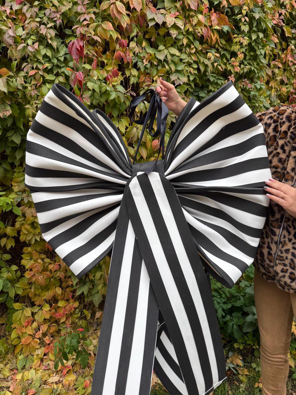 May include: A large, oversized bow with bold black and white stripes. The bow is held up by a black ribbon and appears to be made of a stiff fabric. The background is a lush green and red foliage.