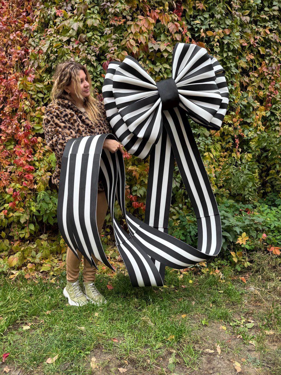 May include: A large, decorative bow with black and white striped ribbon. The bow is held up against a backdrop of green and red foliage. The bow's design features a large, layered bow with long, flowing ribbons.