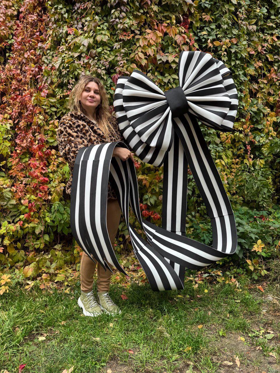 May include: A large, decorative bow with black and white stripes. The bow is held by a person standing in front of a backdrop of green and red foliage. The bow's ribbon hangs down.