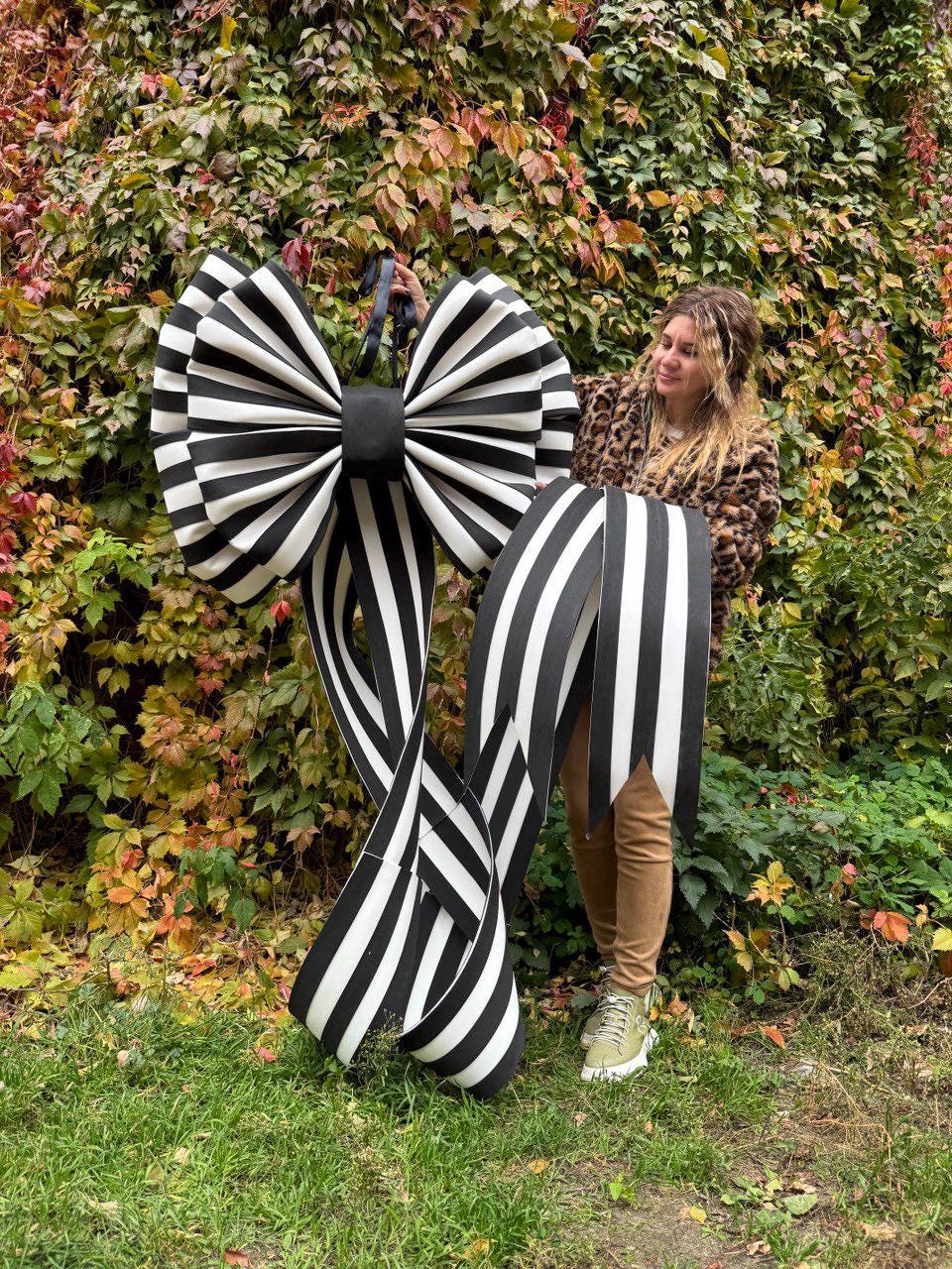 May include: A large black and white striped bow with long, flowing ribbons. The bow features a central black knot and is held up against a backdrop of green and red foliage. The bow is a decorative item.