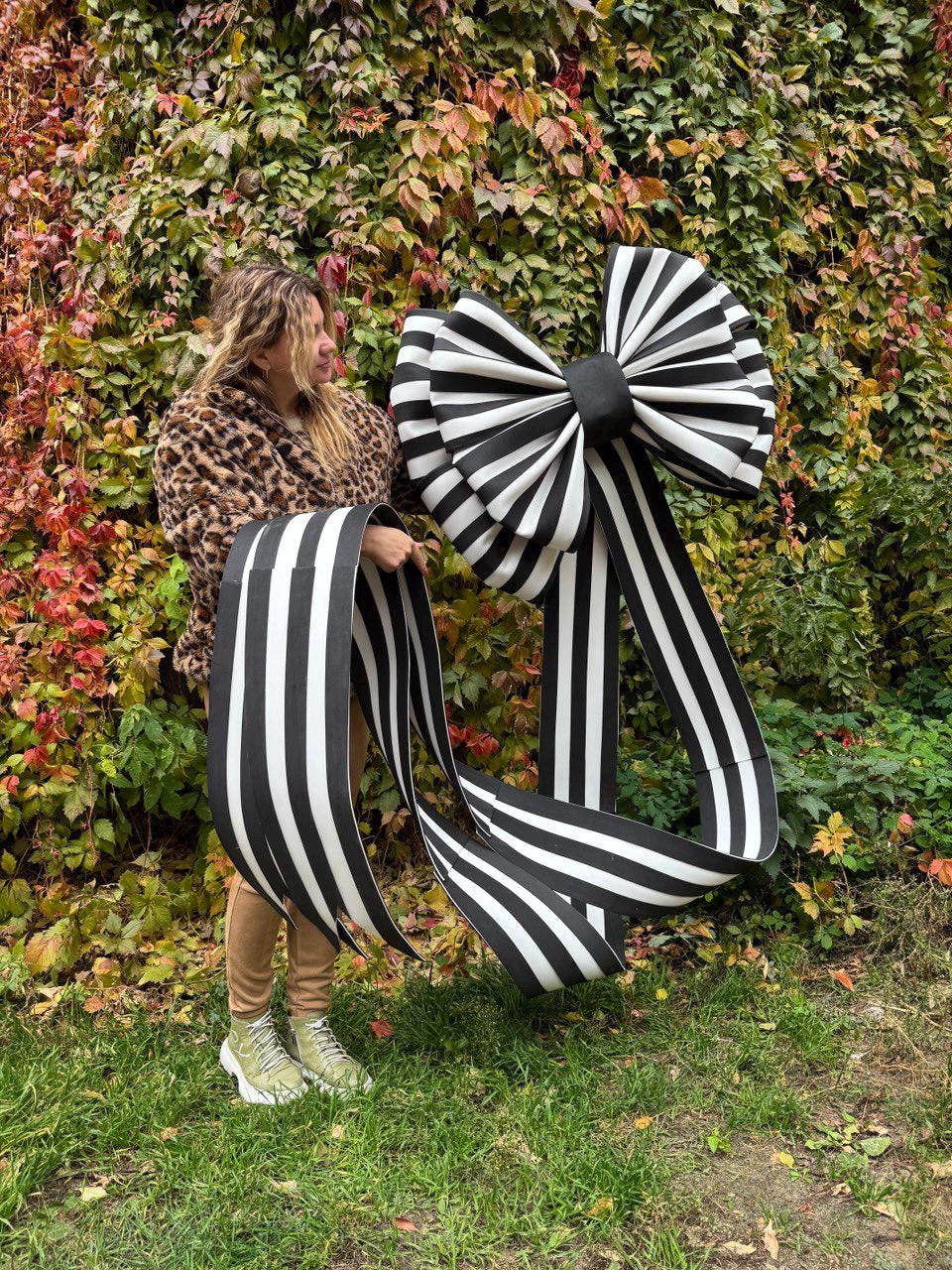 May include: A large, decorative bow with black and white stripes. The bow has a central knot and long, flowing streamers. The bow is held by a person in front of a backdrop of green and red foliage.