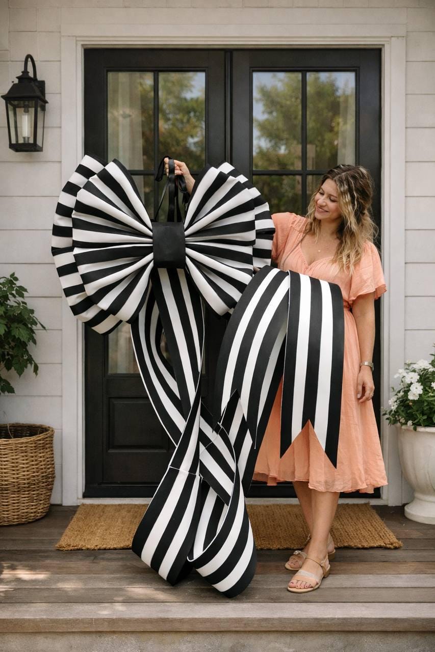 May include: A large, decorative bow with black and white stripes. The bow is held by a person in front of a black door. The bow has long, striped ribbons and a black center.