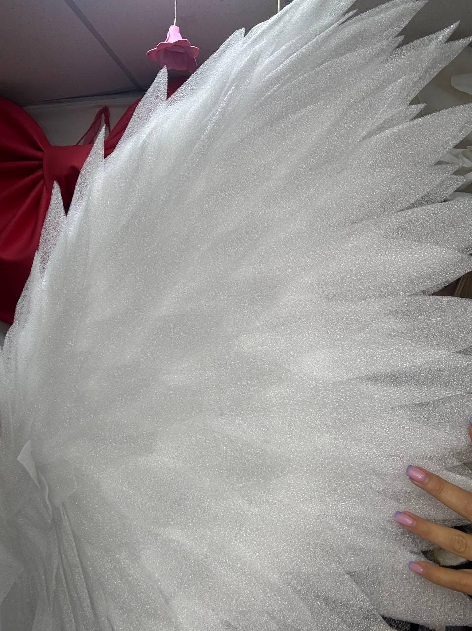 May include: Close-up of a large, white, feathery wing structure. The wing has a layered design with pointed edges, creating a textured appearance. A red bow is visible in the background. The wing is likely a decorative item.