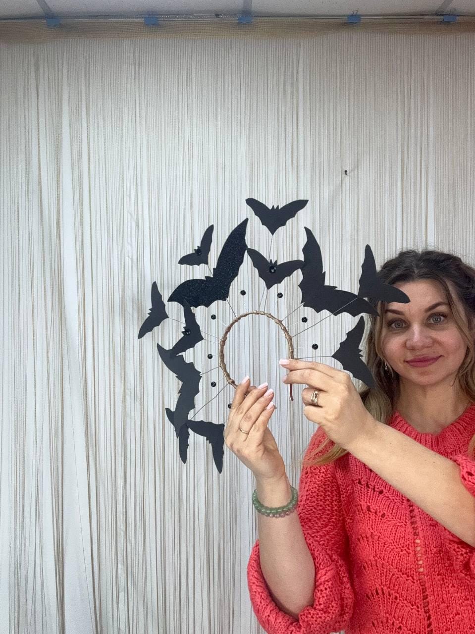 May include: A Halloween-themed wreath featuring black bat silhouettes arranged around a circular frame. The bats vary in size and some have glitter accents. The wreath is held up against a white curtain backdrop.