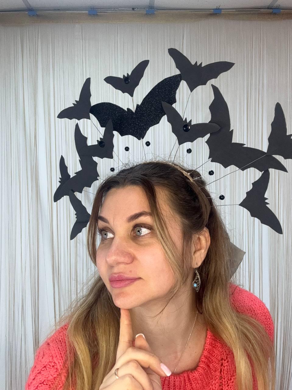 May include: A Halloween-themed headpiece featuring black bat silhouettes. The bats are attached to a thin, gold-colored headband with small black beads. The central bat has a glittery texture. The headpiece is worn by a person.
