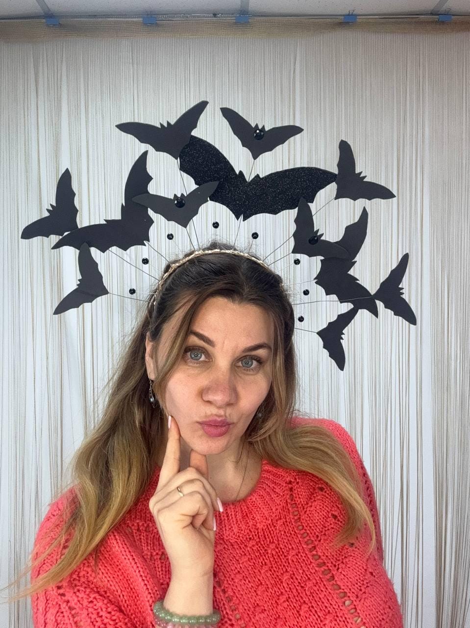 May include: A Halloween-themed headpiece featuring black bat silhouettes. The bats are arranged around a gold-colored headband. The headpiece is worn by a person wearing a red sweater.