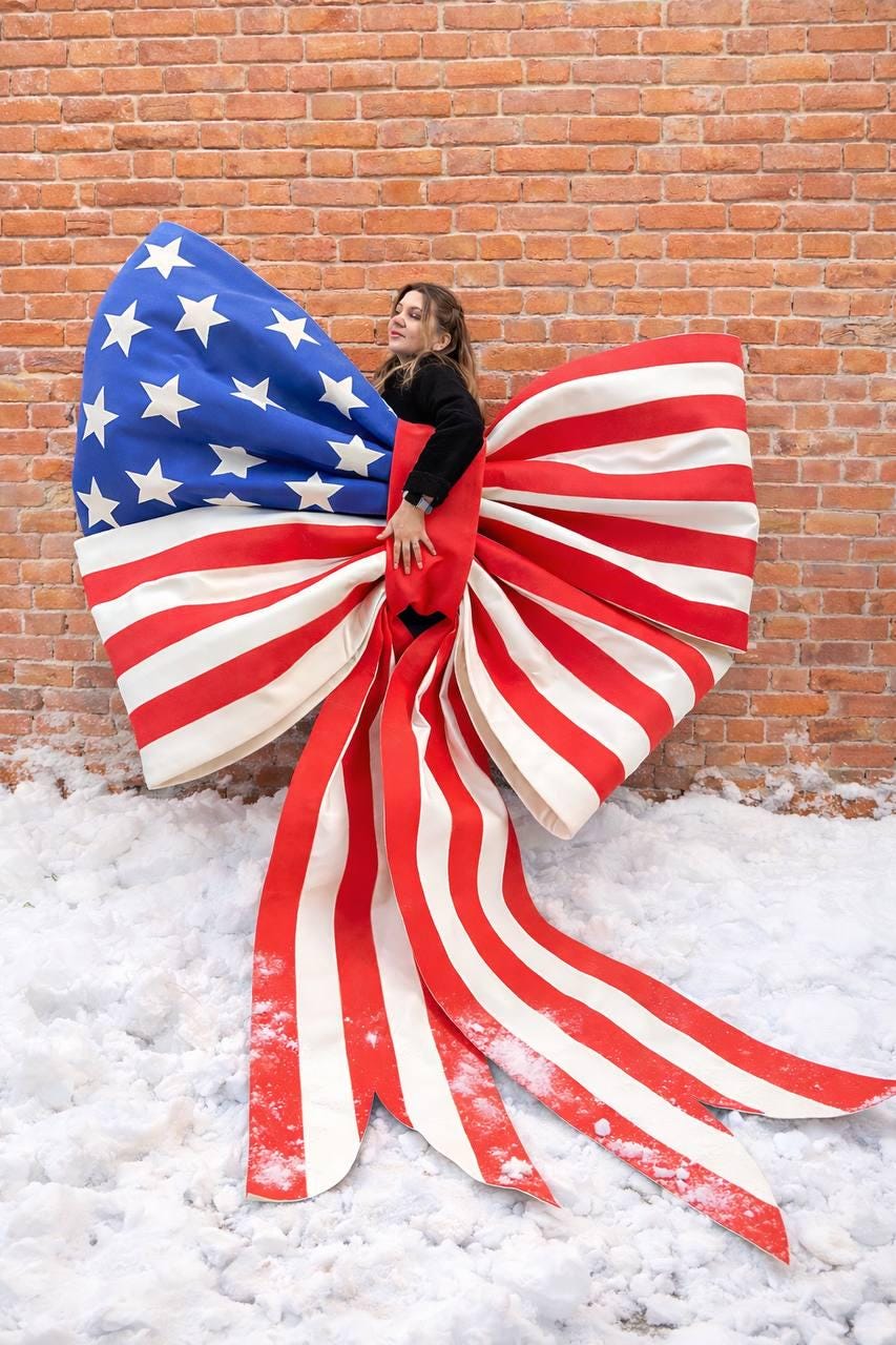 May include: A large bow in the style of the American flag, with red and white stripes and a blue section with white stars. The bow is displayed against a brick wall and snow.