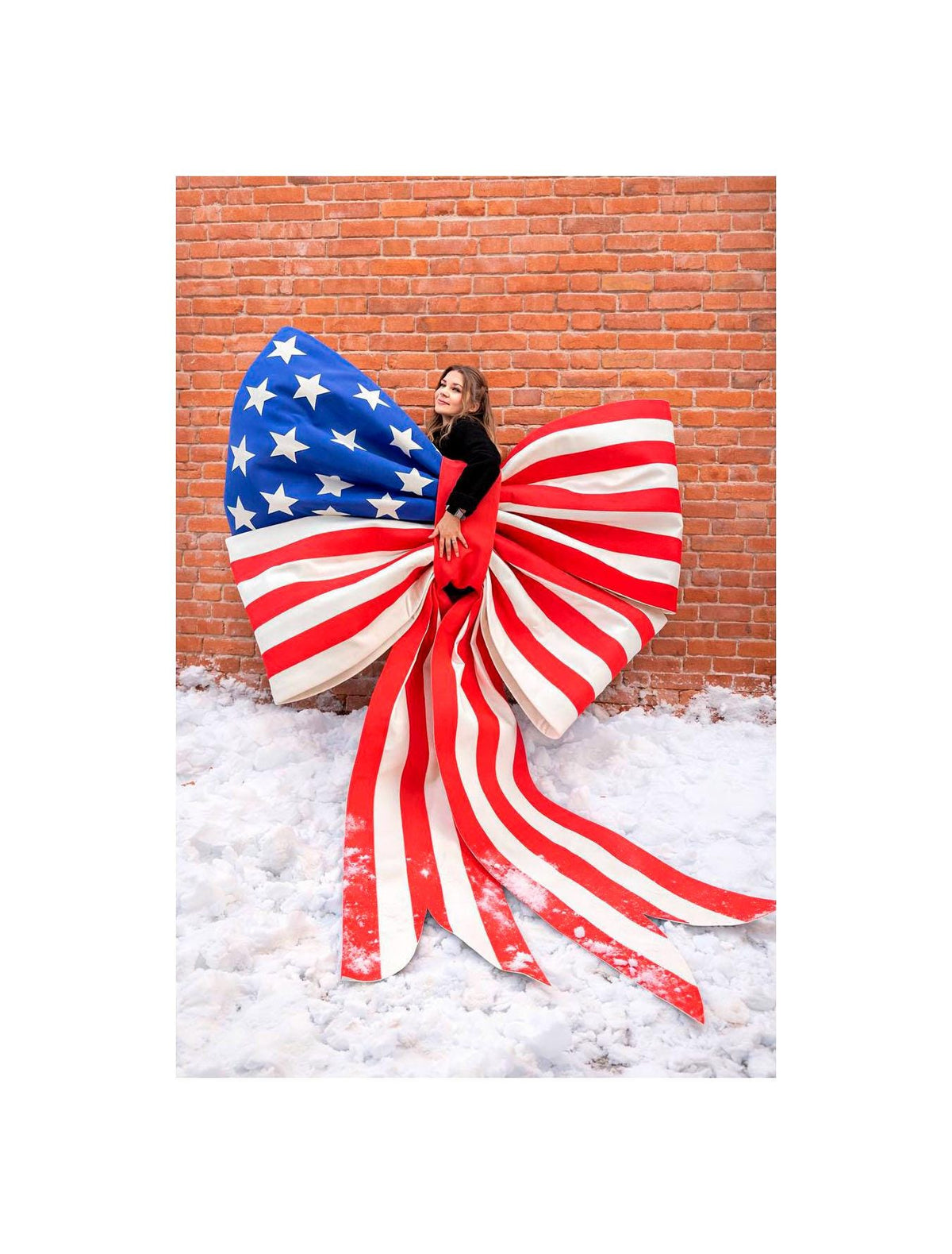 May include: A large decorative bow featuring the American flag design. The bow has a blue section with white stars and red and white stripes. The bow is displayed in front of a brick wall and snow.