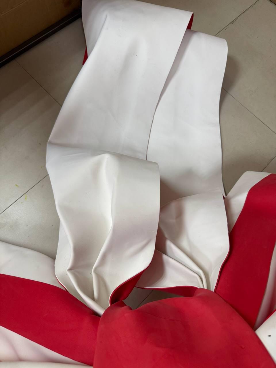 May include: White fabric with red trim is folded and layered. The fabric appears to be thick and possibly waterproof. The image is taken from an overhead angle, showing the folds and curves of the material.