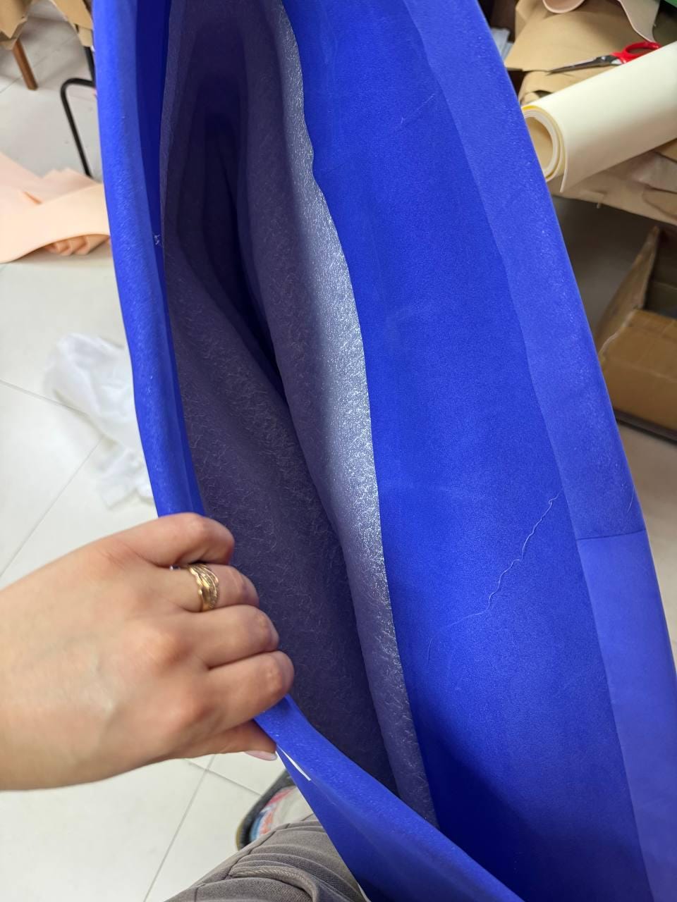 May include: A vibrant blue fabric, possibly a textile or material, is held open, revealing a layered interior. The layers appear to be a light gray, possibly padding or lining. The fabric's texture is smooth and the overall impression is of a folded or wrapped item.