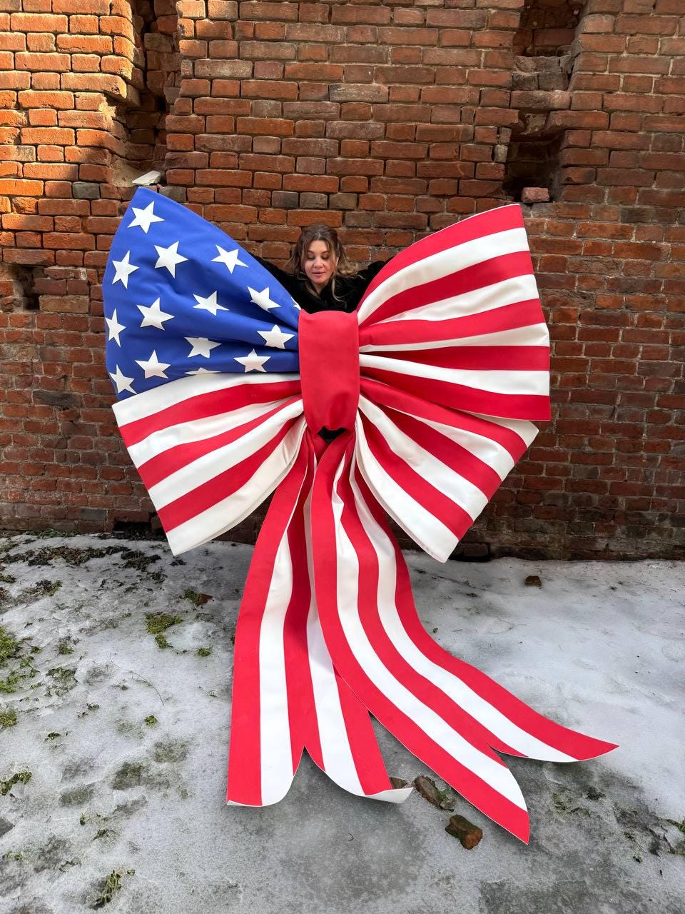 May include: A large decorative bow in the style of the American flag. The bow features a blue section with white stars, and red and white stripes. The bow is made of fabric and is displayed outdoors against a brick wall.