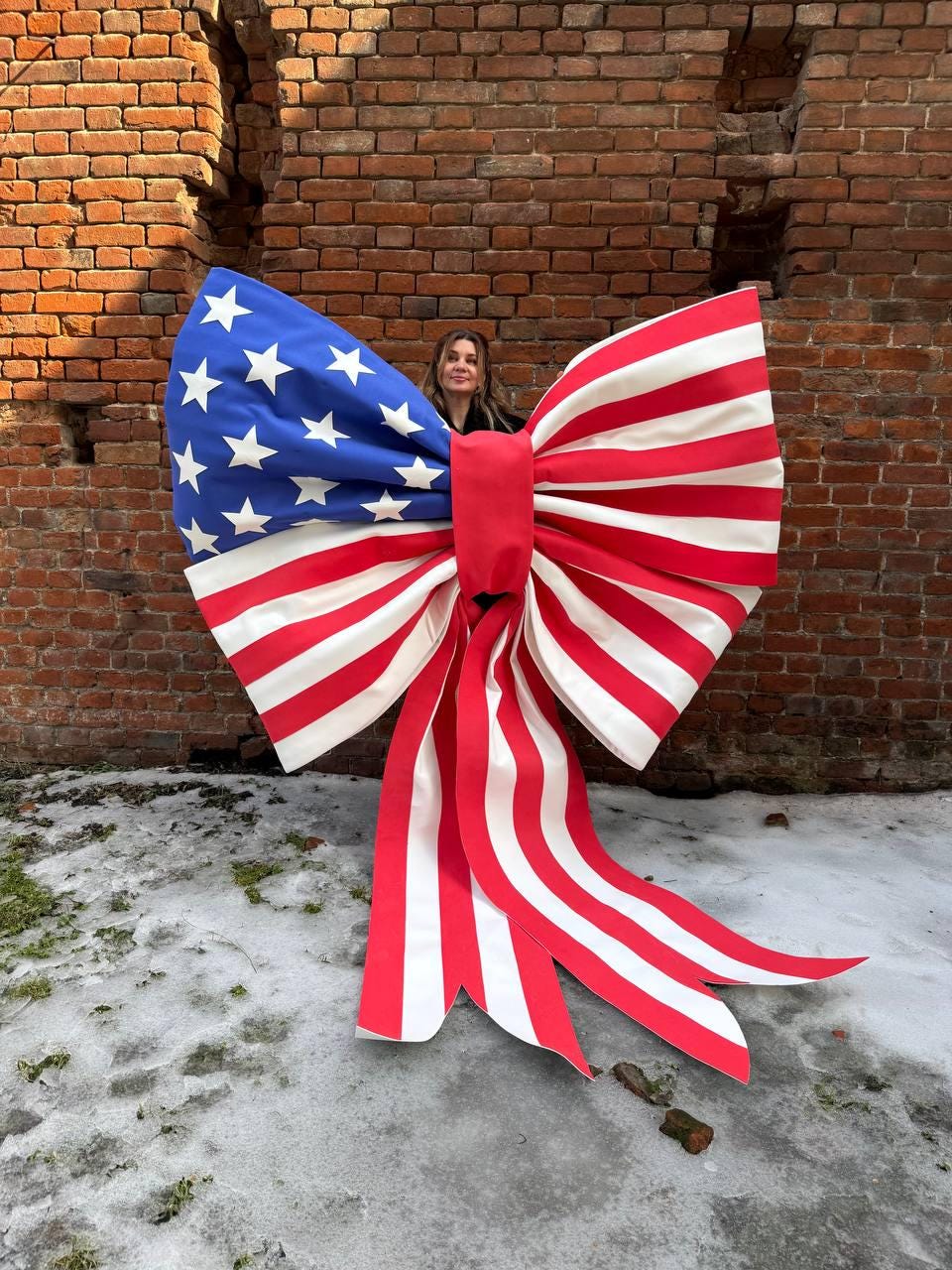 May include: A large decorative bow featuring the American flag design. The bow has a blue section with white stars and red and white stripes. The bow is made of a fabric material and has long, flowing tails.